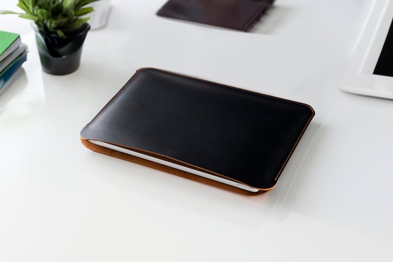May include: A black leather laptop sleeve with brown stitching. The sleeve is closed and lying on a white surface.