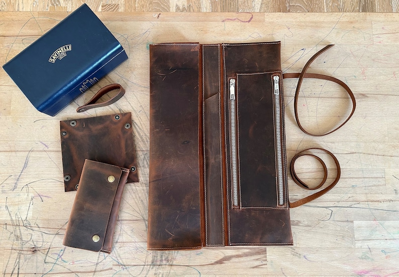 Op de afbeelding: Een bruine leren oprolbaar gereedschapshouder met twee ritsvakken en een lange bruine leren riem. De houder is open en ligt plat op een houten oppervlak. Er zijn twee kleinere bruine leren stukken met klinknagels en een blauwe doos met gouden letters op het oppervlak.