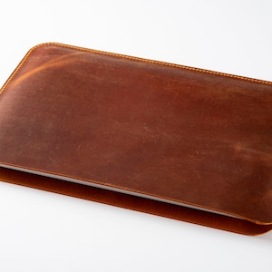May include: A brown leather laptop sleeve with a subtle distressed look. The sleeve has a stitched edge and is closed.