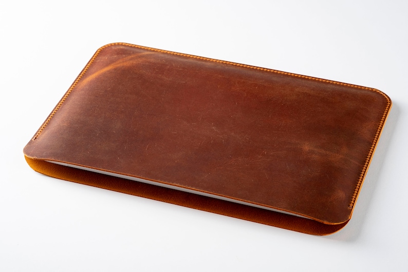 May include: A brown leather laptop sleeve with a subtle distressed look. The sleeve has a stitched edge and is closed.