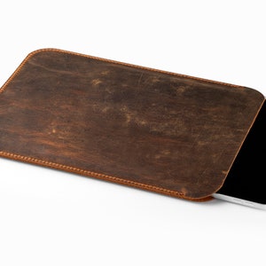 May include: A brown leather tablet sleeve with a stitched border. The sleeve is made of distressed leather and holds a white tablet with a black screen. The sleeve is designed to protect the tablet.