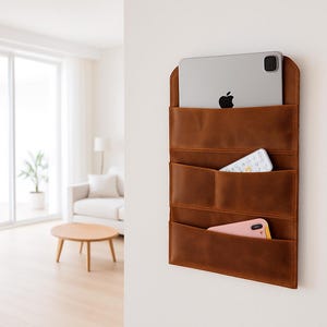 May include: A brown leather wall organizer with multiple pockets, holding a tablet, remote, and phone. The organizer is mounted on a white wall, offering a stylish storage solution for home or office. The design is minimalist and functional.