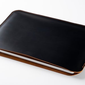 Könnte beinhalten: Eine schwarze Laptop-Hülle aus Leder mit braunem Wildleder-Innenfutter. Die Hülle hat eine genähte Bordüre in einem kontrastierenden orangefarbenen Faden. Der Laptop ist teilweise im Inneren der Hülle sichtbar.