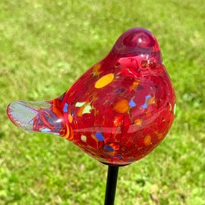 May include: A red glass bird garden stake with colorful flecks, mounted on a black metal pole. The bird has a clear glass wing and a solar panel at the base of the stake, set in a grassy yard.