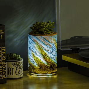 May include: A decorative lamp with a blue, green, and brown swirl pattern. The lamp is lit and casting a warm glow. The lamp is on a wooden surface with a record player and books.