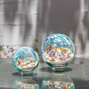 May include: Two decorative glass globes filled with sand and seashells. The globes have a blue and white swirl pattern. The shells are various colors, including pink, orange, and white. The globes sit on a reflective surface.