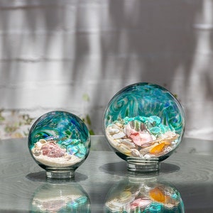 May include: Two glass spheres with a swirling blue and green pattern, filled with sand and seashells. The larger sphere is on the left and the smaller sphere is on the right.