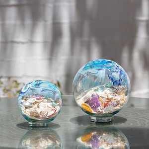 May include: Two clear glass globes filled with sand and seashells. The globes have a blue and white painted design on top. The shells are various colours, including white, purple, and orange. The globes sit on a clear glass base.