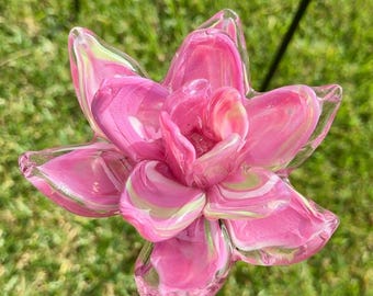 May include: A decorative garden stake featuring a sculpted glass flower with layered petals in shades of vibrant pink, soft white, and pale green. The glass petals have a smooth, glossy finish and are arranged in a multi-layered bloom pattern.