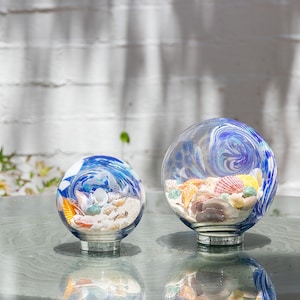 May include: Two glass orbs with blue and white swirl designs, filled with seashells and sand, on a clear glass table.