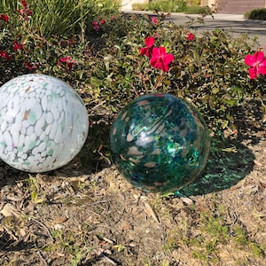 May include: Two decorative glass orbs, one white with green and pink flecks, the other green with white and pink flecks, sitting on a bed of brown mulch.