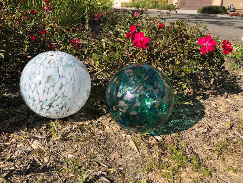 May include: Two decorative glass orbs, one white with green and pink flecks, the other green with white and pink flecks, sitting on a bed of brown mulch.