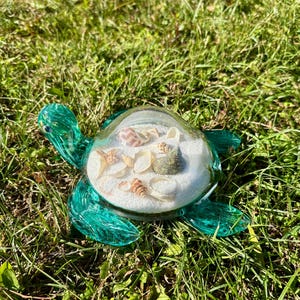 Puede incluir: Una figurita de tortuga de vidrio turquesa con una cúpula de vidrio transparente llena de arena blanca y conchas.