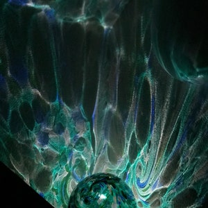 May include: A green glass orb with white and blue swirls and flecks, illuminated from within, casting a swirling pattern of light and shadow on a dark background.