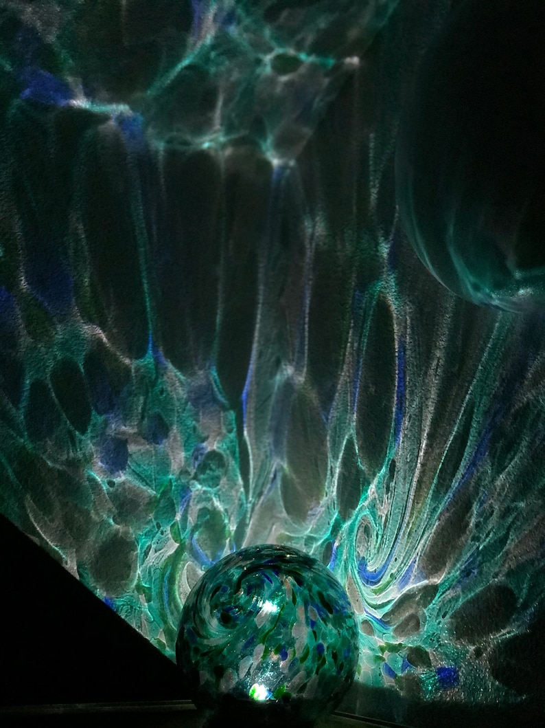 May include: A green glass orb with white and blue swirls and flecks, illuminated from within, casting a swirling pattern of light and shadow on a dark background.
