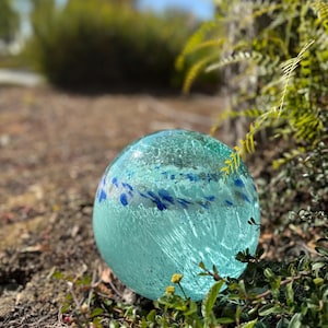 May include: A large, round, light blue glass orb with a crackled texture and blue and white flecks throughout. The orb is sitting on a bed of green foliage.