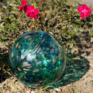 May include: A large, round, teal-colored glass orb with gold flecks and a mottled texture. The orb is sitting on a bed of brown dirt and green grass.