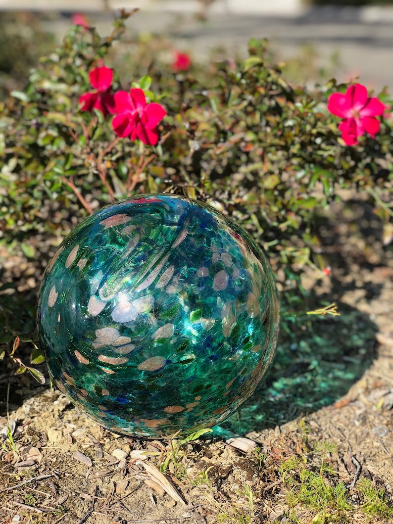 May include: A large, round, teal-colored glass orb with gold flecks and a mottled texture. The orb is sitting on a bed of brown dirt and green grass.
