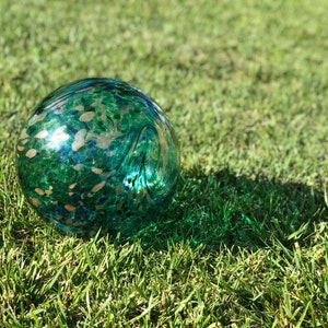 May include: A green glass sphere with gold flecks and blue accents. The sphere is sitting on a bed of green grass.