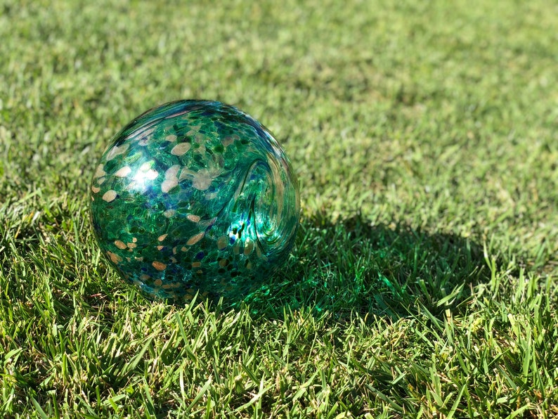 May include: A green glass sphere with gold flecks and blue accents. The sphere is sitting on a bed of green grass.