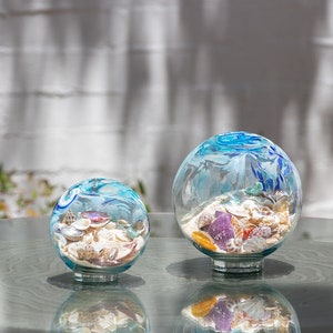 May include: Two glass globes with a blue and white painted design on top, filled with sand and seashells. The globes sit on a clear glass base, reflecting the light. The larger globe is approximately 15 cm in diameter.