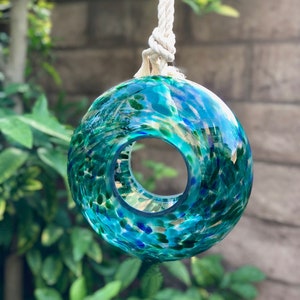 May include: A teal blue glass ornament with a hole in the center, hanging from a white rope. The ornament is covered in small green, blue, and gold flecks.