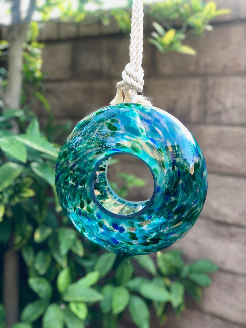 May include: A teal blue glass ornament with a hole in the center, hanging from a white rope. The ornament is covered in small green, blue, and gold flecks.