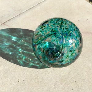 May include: A teal blue glass orb with white and green swirls and speckles. The orb is sitting on a light gray surface.