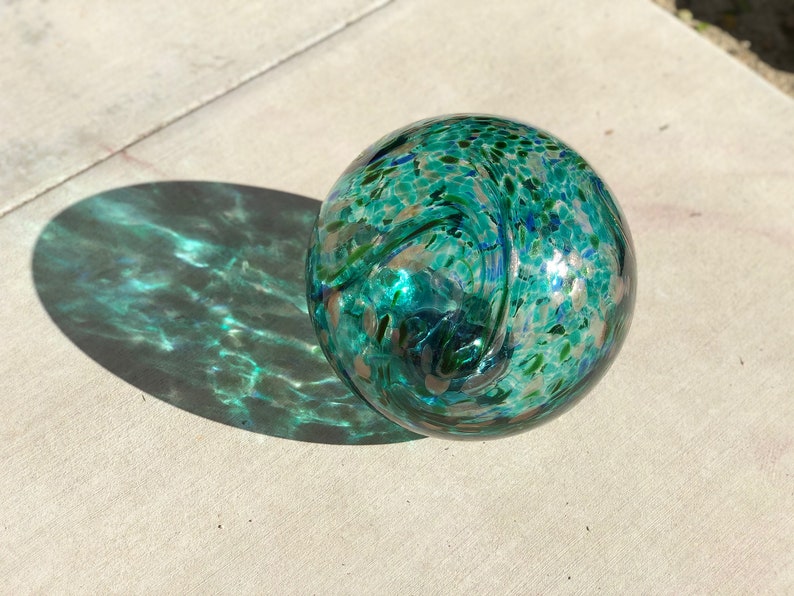 May include: A teal blue glass orb with white and green swirls and speckles. The orb is sitting on a light gray surface.