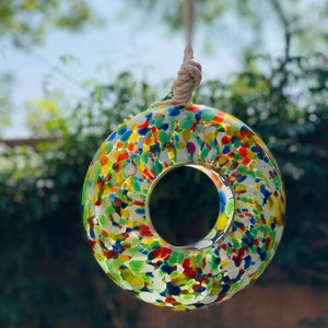 May include: A colorful glass bird feeder with a round shape and a hole in the center. The feeder is suspended by a white rope and is covered in a mosaic of small, colorful glass pieces.