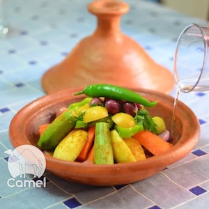Puede incluir: Una tagine de terracota con tapa, llena de una variedad de verduras, incluyendo zanahorias, calabacines, pimientos verdes y patatas. Se vierte agua sobre las verduras desde una jarra de vidrio.