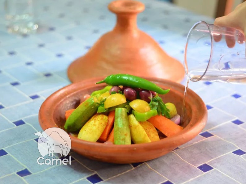 May include: A terracotta tagine with a lid, filled with a variety of vegetables, including carrots, zucchini, green peppers, and potatoes. Water is being poured over the vegetables from a glass pitcher.