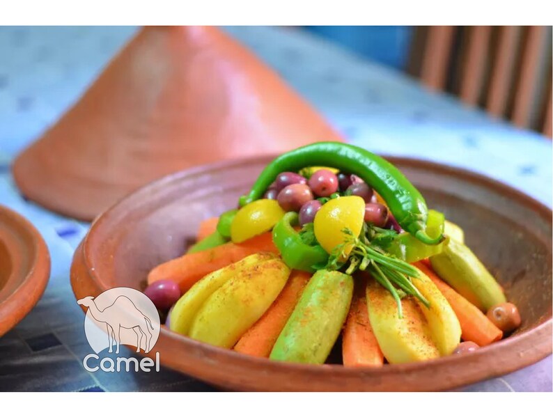 May include: A traditional Moroccan tagine dish with a colorful arrangement of vegetables, including carrots, zucchini, peppers, and olives. The vegetables are arranged in a circular pattern and topped with a green chili pepper. The tagine is served in a terracotta dish with a lid.