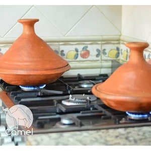May include: Two terracotta tagines, or traditional Moroccan cooking pots, are on a gas stovetop with the flames lit underneath. The tagines are round with a domed lid and are made of a reddish-brown clay.