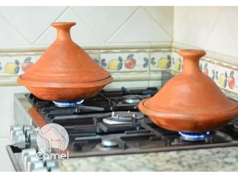 May include: Two terracotta tagines, or traditional Moroccan cooking pots, are on a gas stovetop with the flames lit underneath. The tagines are round with a domed lid and are made of a reddish-brown clay.