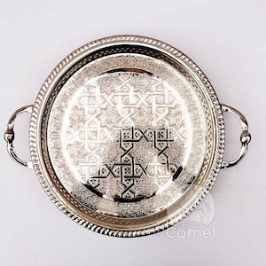 Peut inclure: Un plateau de service rond en argent avec des motifs géométriques ornés et deux poignées. Le plateau est rond et possède un bord surélevé.
