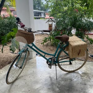 Könnte beinhalten: Ein blaues Fahrrad mit einem Weidenkorb vorne und einem Weidenkorb hinten. Das Fahrrad steht auf einer Betonfläche.