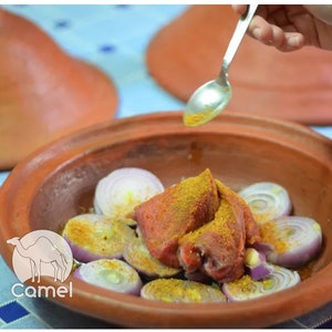 May include: A close-up of a clay tagine with red meat, onions, and spices. The meat is covered in a yellow spice blend. A hand is pouring oil from a spoon over the meat.