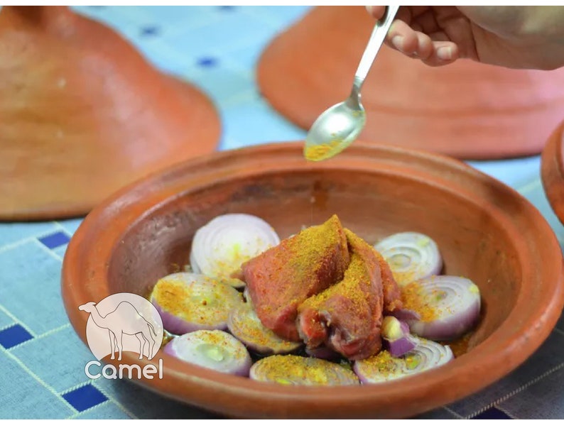 May include: A close-up of a clay tagine with red meat, onions, and spices. The meat is covered in a yellow spice blend. A hand is pouring oil from a spoon over the meat.