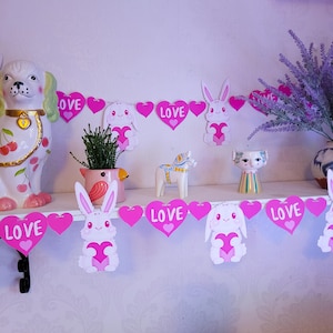 May include: Pink and white heart-shaped banner with the word "LOVE" on each heart. The banner is decorated with white bunnies holding pink hearts.