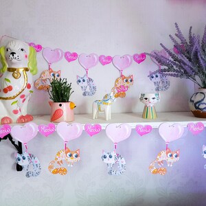 May include: A pink and white garland with pink hearts and cute cartoon cats holding balloons that say "Meow".