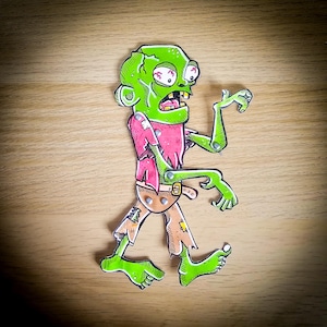 Può includere: Illustrazione di uno zombie verde con occhi rossi, una camicia rosa e pantaloni color camoscio strappati. Lo zombie ha giunture articolate e un'espressione sorpresa. L'opera d'arte è su una superficie di legno.