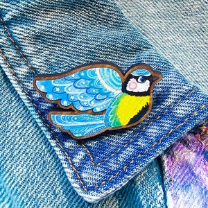 May include: A wooden bird pin with a blue, yellow, and white design. The pin is in the shape of a bird with its wings spread. The pin is attached to a denim jacket.