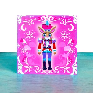 Puede incluir: Una caja decorativa cuadrada rosa con un conejo de dibujos animados vestido de cascanueces. El conejo tiene pelaje marrón, una cara azul y blanca y viste un uniforme rojo y azul. El fondo es rosa con diseños florales y de copos de nieve blancos.