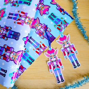 May include: Gift wrap and gift tags featuring a whimsical nutcracker rabbit design. The wrapping paper has a light blue background with repeating illustrations of the rabbit nutcracker and snowflakes. The gift tags are cut-out shapes of the rabbit nutcracker.