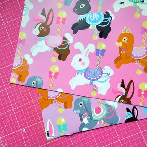 May include: Pink wrapping paper with a repeating pattern of cartoon bunnies wearing colourful dresses and riding carousel horses. The bunnies are pink, white, and brown. The horses are white, brown, and black. The background is pink with white polka dots.