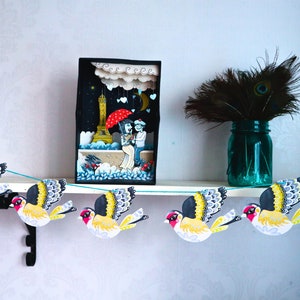 May include: A paper garland featuring colourful birds, each with a yellow body, black wings, and a red beak. The garland is strung on a blue string and is hanging from a shelf. There is a small wooden box with a scene of a couple in front of the Eiffel Tower on the shelf.
