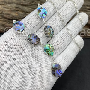 May include: A silver necklace with iridescent abalone shell pendants in oval and teardrop forms. The abalone shells show blue, green, and purple hues. The necklace is displayed on a white surface.