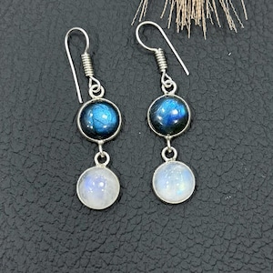 May include: A pair of silver earrings with a two-stone design. Each earring features a round, iridescent blue stone at the top and a round, white stone below. The earrings have a simple hook closure.