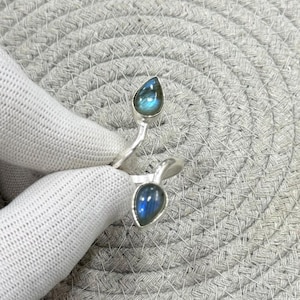 May include: A silver ring with two teardrop-shaped, iridescent blue gemstones. The ring's design has a split band that curves around the finger, with a gemstone at each end. The gemstones have a polished finish.